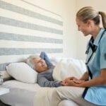 Friendly nurse with senior woman at home. Smiling nurse talking with her patient while getting her out of bed. Disabled woman getting regular health checkup done at home with doctor.